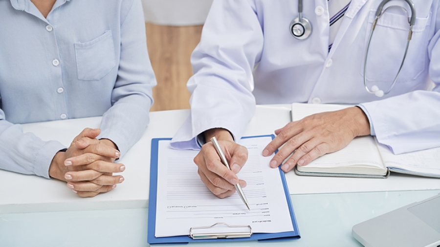 Close-up shot of male doctor wearing white coat filling in medical history while sitting at desk next to female patient